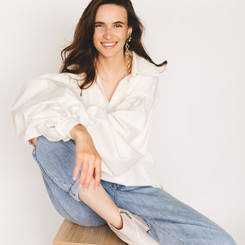 A woman sitting on a wooden stool wearing a white oversized blouse with dramatic puffed sleeves, light blue jeans, and white ankle boots, against a plain white background.